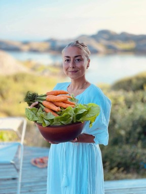 Kvinne i blå kjole holder en bolle med salat og gulrøtter hun selv har dyrket. I bakgrunnen ser vi et kystlandskap.