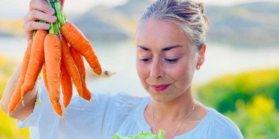 Ung kvinne i blå kjole holder en bunt med gulrøtter i hånden. I den andre hånden har hun en stor bolle med frisk salat. Kvinnen ser ned på salaten. - Foto: Gilberto Debelian